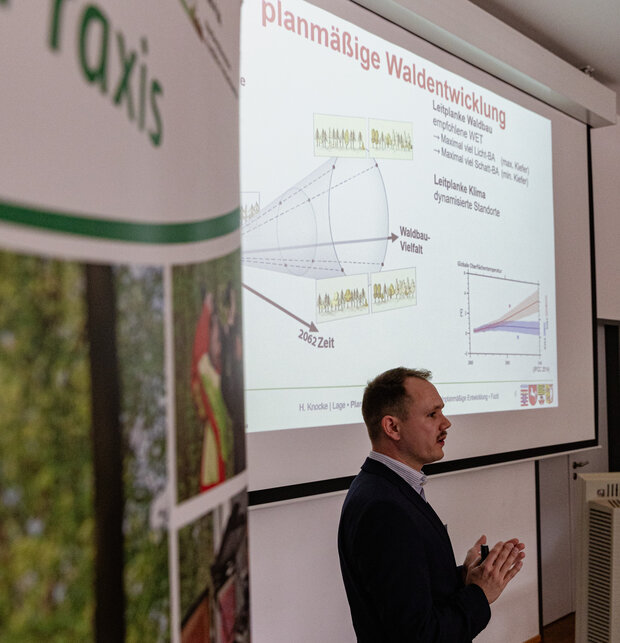 Redner steht vor einer Leinwand mit Vortragsfolien.